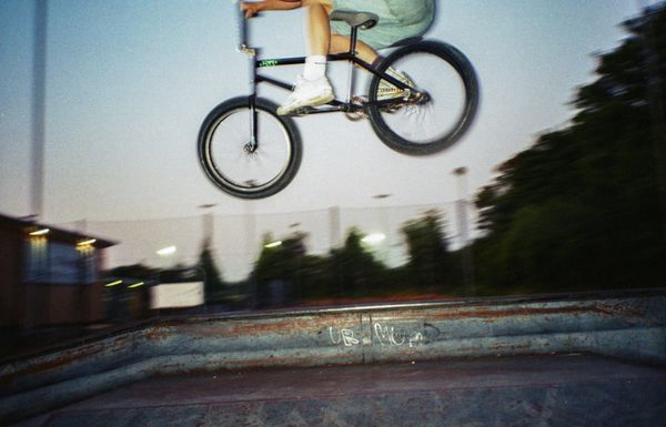 Titled Aerial Momentum, this photograph captures the thrill and freedom of BMX riding. The rider is suspended mid-air, bike tilted forward, frozen in a moment of gravity-defying action. Shot with a slight motion blur, the image conveys the speed and energy of the jump, creating an almost dreamlike sense of flight. The dusk lighting adds a sense of mystery, with only the faint glow of nearby lights and blurred greenery in the background. This image encapsulates the raw, uninhibited spirit of BMX culture, celebrating the courage and skill required to take on the ramps and the air.